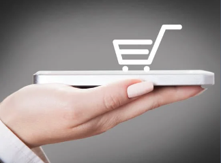Imagem: Canva A imagem mostra uma mão, de pele clara e unhas pintadas de um tom neutro segurando horizontalmente um smartphone ou tablet prateado. Flutuando acima da tela do dispositivo está um ícone branco estilizado de um carrinho de compras, como os usados para compras online (e-commerce). O fundo é um tom sólido e neutro de cinza escuro.