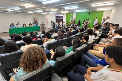A imagem mostra um auditório cheio, com dezenas de pessoas sentadas assistindo a um evento. Na frente da sala, há uma mesa coberta com uma toalha verde, onde estão sentados seis integrantes da mesa de abertura. Ao lado direito, em pé, há um grupo de pessoas aguardando ou acompanhando a cerimônia. As paredes do auditório são claras, e ao fundo há cortinas verdes e bandeiras posicionadas junto à mesa principal.