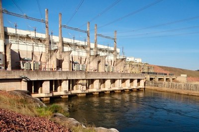 Usina Hidrelétrica Colíder, no rio Teles Pires (MT). Foto: Aneel Fotografia da Usina Hidrelétrica Colíder no Rio Teles Pires, em Mato Grosso, mostrando a barragem de concreto e o vertudouro no centro. A estrutura divide a paisagem: de um lado, a água calma e verde-azulada do reservatório; do outro, o rio continua o curso, com o leito parcialmente seco e áreas de terra batida visíveis. A usina está cercada por uma densa vegetação verde de mata atlântica nas margens, sob um céu claro com poucas nuvens.