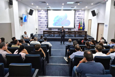 Ambiente interno de auditório moderno, com várias pessoas sentadas em cadeiras azuis acolchoadas, voltadas para um palco. No palco, há um palestrante em pé, falando diante de uma tela grande.
