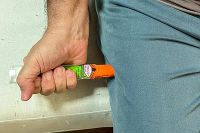 Caneta de adrenalina. Foto: Fiocruz. Fotografia da mão de um homem aplicando uma caneta de adrenalina em sua própria perna, vestido com uma calça azul.