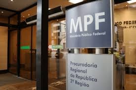 Fonte: Ascom/MPF 2a Região imagem em plano fechado mostra a sinalização de entrada de um edifício. Em primeiro plano, uma placa cinza e azul exibe a sigla MPF e abaixo, em letras menores, Ministério Público Federal. Abaixo desta, uma segunda placa indica Procuradoria Regional da República 2ª Região. Ao fundo e à esquerda, uma porta de vidro da entrada do prédio é visível.