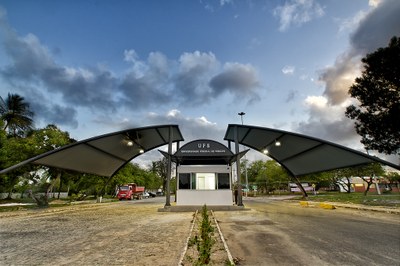 Imagem: Adilson Andrade/Ascom UFS Entrada principal da Universidade Federal de Sergipe, com guarita central e duas coberturas metálicas curvas em destaque sob um céu parcialmente nublado.