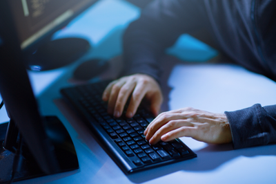 Foto ilustrativa: Canva Close-up das mãos de uma pessoa digitando em um teclado preto em uma mesa escura, iluminada por uma luz azulada vinda da tela de um monitor, sugerindo que a pessoa está trabalhando no computador em um ambiente escuro (possivelmente programação ou cibercrime).