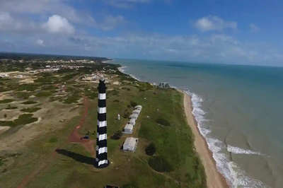 Vista aérea do Farol do Calcanhar em Touros, Rio Grande do Norte, uma estrutura alta e cilíndrica listrada em preto e branco, em uma área de vegetação rasteira ao lado de uma praia de areia. O mar turquesa se estende sob um céu azul com nuvens. Um pequeno núcleo urbano é visível ao longe, à esquerda.