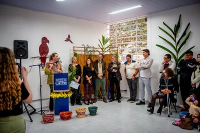 Fotografia colorida em ambiente interno, mostrando um grupo de seis pessoas, quatro homens e duas mulheres, em pé e lado a lado. Quatro pessoas estão na frente, incluindo um homem de cabelo grisalho no centro que usa óculos e terno escuro, e uma mulher à sua direita com uma blusa escura e um colar colorido de miçangas. Atrás deles, há duas mulheres, uma delas à esquerda, sorrindo e usando uma blusa bege e um cocar indígena. Todos olham para a câmera em frente a uma parede de tom pastel. A imagem registra um momento da inauguração da Casa do Estudante Indígena da UFPR.
