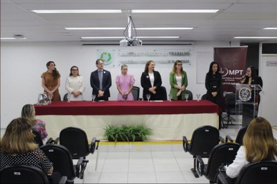 Foto: Ascom MPT-PB Sete pessoas estão sentadas atrás de uma mesa longa coberta com toalha vermelha e bege, em um evento formal do Ministério Público do Trabalho. Há uma placa na parede com o nome da instituição e uma planta decorativa à frente da mesa