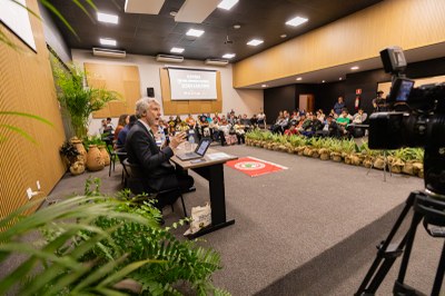 Fotos: Leobark Rodrigues/Comunicação/MPF Foto de uma sala moderna e bem iluminada. Um palestrante está sentado à frente com microfone e laptop, falando para uma plateia atenta. Há plantas decorativas, vasos grandes e uma câmera gravando o evento.