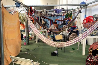 Foto: Catarina Barbosa/Brasil de Fato Foto de abrigo improvisado com armação metálica, redes, roupas penduradas e pertences, com uma pessoa sentada ao centro.