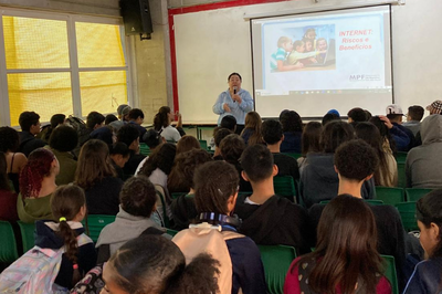 Foto: Ascom/PRR3 A imagem mostra uma palestra sendo ministrada para um grupo de estudantes em um pátio. A palestrante está em pé, segurando um microfone, enquanto apresenta um slide projetado na tela com o título “INTERNET: Riscos e Benefícios”. Os alunos estão sentados em cadeiras verdes, atentos à apresentação.