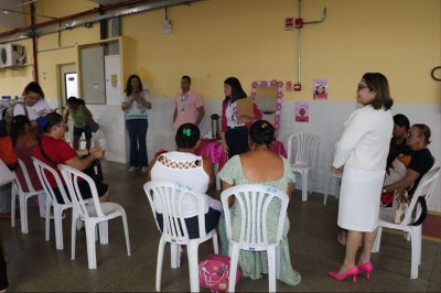 A imagem mostra um grupo de pessoas reunidas em uma sala. Algumas estão sentadas em cadeiras plásticas brancas dispostas em semicírculo, enquanto outras estão em pé. Há uma mesa coberta com uma toalha rosa. Ao fundo, há decorações e cartazes em tons de rosa, com motivos florais, voltados à saúde e valorização da mulher.