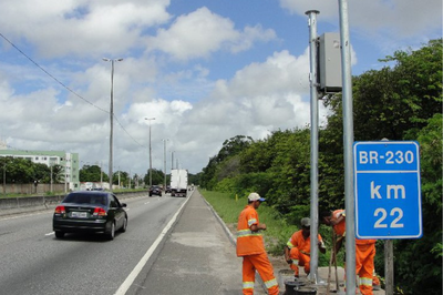 Foto:Dnit/Paraíba Homens usando uniformes laranja de manutenção trabalham na lateral de uma rodovia movimentada, a BR-230, no quilômetro 22, conforme sinalização visível no poste ao lado de um equipamento. Veículos, incluindo um caminhão, circulam na pista.