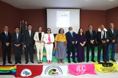 5ª Conferência Estadual dos Direitos Humanos da Paraíba, em João Pessoa, em 9/10/2025. Foto: Comunicação/MPF. Fotografia colorida em ambiente interno de um evento ou conferência, focada em quatro pessoas, aparentemente representantes institucionais, em frente a um painel de fundo. No primeiro plano, à esquerda, um homem com camisa social branca e gravata escura, com um crachá pendurado, segura um microfone com a mão esquerda, enquanto gesticula com a direita. Ao seu lado, três mulheres estão sentadas, com microfones à frente. A primeira mulher sorri e veste uma roupa formal escura, com o crachá visível. O painel de fundo atrás delas exibe a logomarca do MPF - Ministério Público Federal, o slogan "Direitos Humanos, o Brasil que a gente quer" e a identificação do evento: "5ª Conferência Estadual de Direitos Humanos". O ambiente está bem iluminado, sugerindo a realização de um debate ou mesa de discussão.