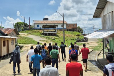 MPF visita a comunidade indígena de Feijoal, em Benjamin Constant (AM), em 17/9/2025. Foto: Comunicação/MPF. Esta imagem mostra um grupo de aproximadamente 15 jovens e homens caminhando por uma rua de terra e cimento em uma área que parece ser rural ou uma pequena comunidade, possivelmente na Amazônia ou em uma região de clima tropical. Eles estão se afastando da câmera, em direção a um terreno mais alto onde se destacam algumas construções maiores e antigas, incluindo uma com telhado azul e branco e uma estrutura de madeira ao lado, sob um céu parcialmente nublado e ensolarado. A rua é ladeada por casas simples de madeira com telhados de chapa de metal em ambos os lados, e há postes de eletricidade e vegetação exuberante ao redor, sugerindo um ambiente simples e de vida comunitária.