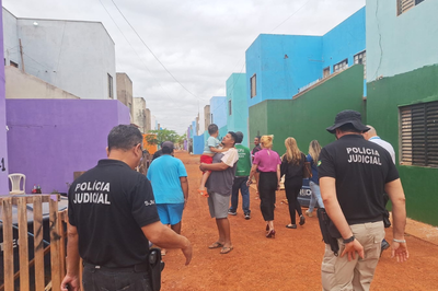 Fotos: Comissão Regional de Soluções Fundiárias do Poder Judiciário de Mato Grosso Agentes da Polícia Judicial e uma equipe de pessoas (civis) caminham por uma rua de terra vermelha em uma área residencial caracterizada por casas geminadas coloridas (azul, verde, roxo e branco). No centro, uma pessoa carrega uma criança ao colo, e os agentes da Polícia Judicial estão em primeiro plano.