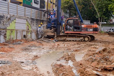 Obras na Avenida Santo Antônio, no Centro, em Garanhuns (PE). Foto: Hilton Marques/Prefeitura de Garanhuns. Fotografia de uma avenida em obras, com as faixas cobertas de lama, construções ao fundo e uma escavadeira sobre a lama, com operários trabalhando nela.