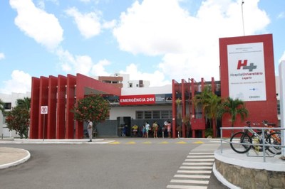 Fotografia da fachada do Hospital Universitário de Lagarto. O prédio possui detalhes em vermelho e parte da fachada na cor branca. Há bicicletas estacionadas à direita e pessoas em pé, na frente da unidade hospitalar.