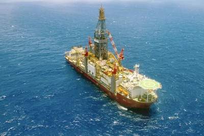 Vista aérea de uma plataforma de perfuração (navio-sonda ou drillship) flutuando em águas azuis profundas no oceano. A plataforma, grande e de cores brancas e vermelhas, possui uma torre de perfuração central e um heliponto na popa.