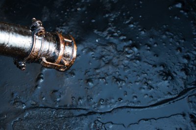 Foto ilustrativa: Anan Kaewkhammul, via Canva. Foto retangular em close-up de um bocal de mangueira metálico coberto por petróleo, que forma uma poça espessa e irregular sobre uma superfície escura abaixo.