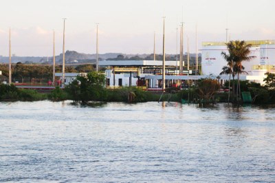 Vista de um corpo d'água, com vegetação na margem, e ao fundo, instalações industriais brancas com postes altos e um coqueiro.