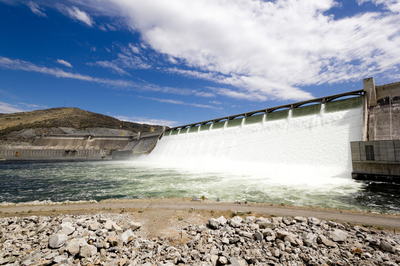 Uma visão ampla e em perspectiva de uma grande barragem hidrelétrica de concreto liberando um volume massivo e branco de água pela sua estrutura, criando turbulência no rio abaixo. Há uma encosta de montanha seca à esquerda e o céu é azul com nuvens brancas.