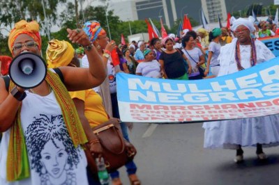 Foto: Marcha das Mulheres Negras/divulgação Foto colorida de uma marcha com mulheres negras. Uma mulher segura um megafone. Outras carregam uma faixa azul com texto em alusão à marcha.