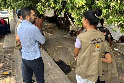 Foto: MPF/RR Em uma calçada arborizada, quatro pessoas estão reunidas conversando, voltadas para a esquerda da imagem. Duas mulheres usam coletes bege com o logotipo da Fundação Nacional dos Povos Indígenas (FUNAI) e a inscrição "Ministério dos Povos Indígenas" nas costas. Um homem de camisa azul clara e calça escura está de costas, conversando com o grupo. Ao fundo, sob as árvores, há redes armadas e pessoas deitadas ou sentadas. Um veículo azul com a porta aberta está estacionado à esquerda. O ambiente sugere uma visita técnica ou ação social em apoio a povos indígenas.