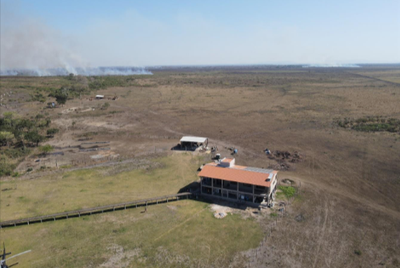 Foto de uma casa no meio de um grande descampado. Ao fundo, há uma queimada na vegetação.