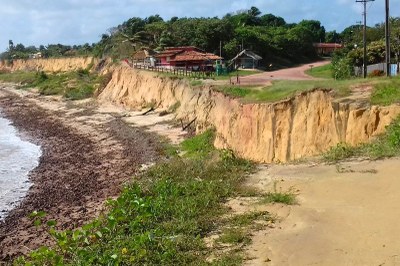 Foto: MPF Foto de uma área de erosão costeira em Maracanã, Pará, com falésias de terra amarelada, vegetação e construções no topo, e mar na esquerda.