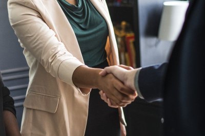 Foto de aperto de mãos entre duas mãos de homens brancos que usam ternos: um cinza e um azul. Ambas as camisas sociais são brancas.