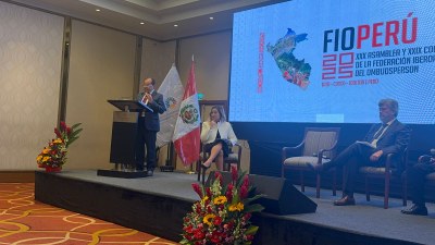 Foto: MPF Foto do procurador federal dos Direitos do Cidadão, Nicolao Dino, no XXIX Congresso e Assembleia Geral Ordinária da Federação Iberoamericana de Ombudsperson (FIO). Ele está em pé em um púlpito. Há outras duas pessoas sentadas no palco.
