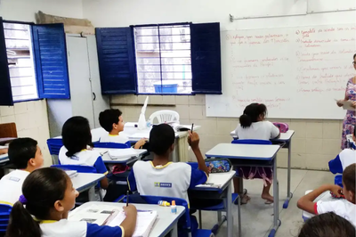 Uma cena de sala de aula de ensino fundamental, com alunos sentados em carteiras azuis e brancas, voltados para um quadro branco onde há perguntas escritas à mão. Uma professora está à direita (apenas parcialmente visível) e um dos alunos levanta a mão para responder uma pergunta.
