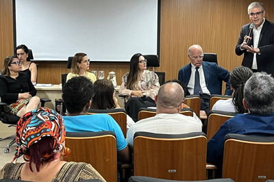 Fotos: Comunicação MPF Uma reunião em uma sala, com um homem de terno e óculos está de pé à direita, falando ao microfone para um auditório sentado. Na mesa, há quatro mulheres e um homem. Em primeiro plano, uma pessoa da plateia usa um turbante colorido.