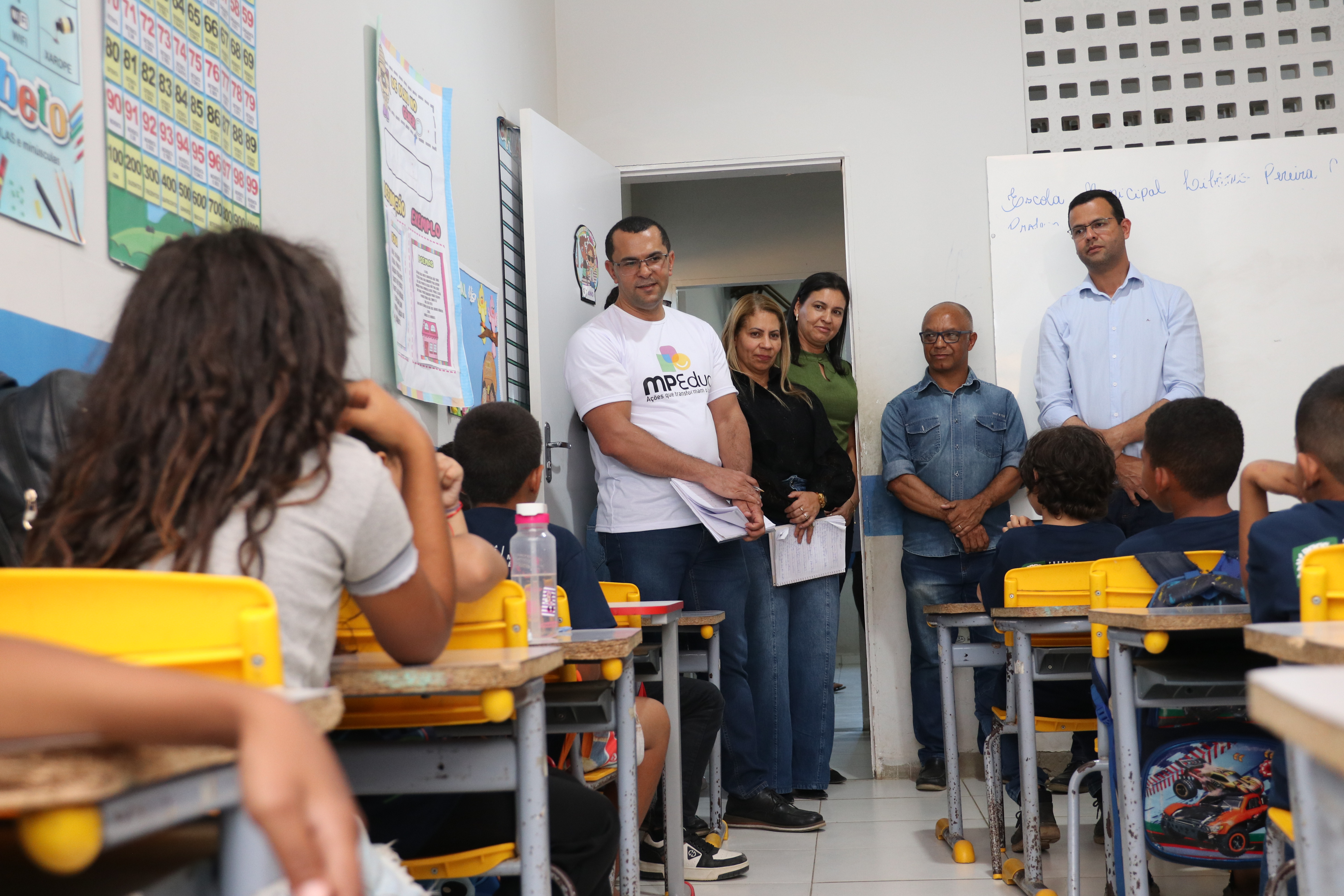 Sala de aula com alunos sentados em carteiras amarelas, de frente para o quadro. Na porta e ao lado, cinco adultos em pé observam a turma, entre eles um homem com camiseta do MPEduc e outros segurando anotações. As crianças estão voltadas para os visitantes, que acompanham a atividade escolar.