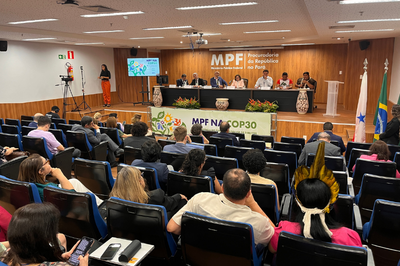Fotos: Comunicação MPF Uma vista de um auditório lotado onde está ocorrendo um evento do MPF na COP30 (Ministério Público Federal). No palco elevado, um painel de sete autoridades está sentado em uma longa mesa, com um grande logo do MPF ao fundo. Uma pessoa, possivelmente indígena, na plateia, usa um cocar amarelo em primeiro plano.