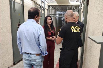 Visita ao presídio Na foto, tirada no interior do presídio, aparecem grades e celas ao fundo e nas laterais. Quatro pessoas estão em primeiro plano, em pé em um corredor estreito. Dentre elas, a procuradora da República Silvia Regina Pontes Lopes, que veste um macacão vermelho, dois conselheiros e um policial penal.