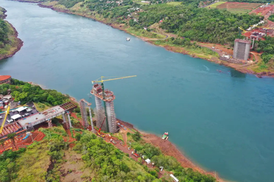 Imagem aérea de um rio largo e de cor azulada, cercado por planícies. Nas margens do rio, há pilares de concreto ainda em fase de construção, guindastes e canteiros de obras