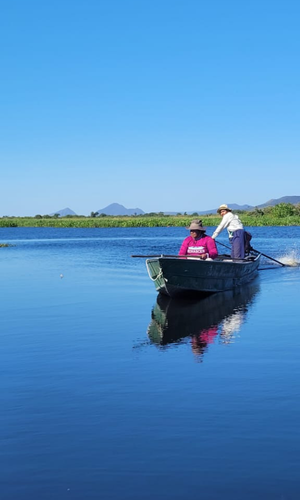 A imagem é um plano vertical mostrando duas pessoas em um pequeno barco a motor navegando em um corpo d'água calmo e azul. O céu é de um azul claro e sem nuvens. Ao fundo, há uma margem coberta de vegetação e, mais distante, pequenas colinas ou elevações.
