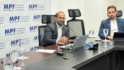 Nesta imagem, dois homens aparecem em destaque, sentados à mesa da sala de reuniões do MPF/AL. O homem à esquerda fala ao microfone, com um notebook e papéis à sua frente, enquanto o outro observa atentamente. Ao fundo, o painel institucional com o logotipo do MPF indica que o encontro é oficial. Copos de água e xícaras completam a mesa, reforçando o caráter formal da reunião.