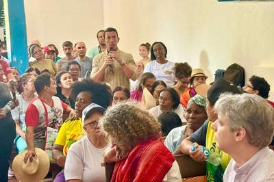 foto mostra um homem de pé, falando ao microfone, dentro da capela, enquanto outras pessoas sentadas o escutam