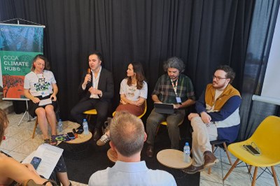 Foto: Divulgação/ CCCA Cinco pessoas participam de um painel de debate em um ambiente interno, sentadas lado a lado em cadeiras amarelas diante de uma cortina preta. Uma delas fala ao microfone enquanto as outras escutam atentamente. À esquerda, há um banner com os dizeres “CCCA Climate Hub”. Na frente, o público acompanha a conversa.