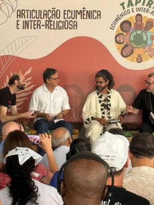 Fotografia em ambiente interno de um evento sobre "ARTICULAÇÃO ECUMÊNICA E INTER-RELIGIOSA". Quatro homens estão sentados em um palco baixo, engajados em um debate. À direita, um homem afrodescendente, com dreads presos em um turbante e usando uma túnica branca com detalhes pretos, segura um papel e gesticula enquanto fala com um homem de camisa social branca e óculos. À esquerda, outro homem com barba veste uma camiseta preta, e um quarto homem, à direita extrema, usa uma camisa escura. O público, com diversas etnias, é visto de costas em primeiro plano, alguns gravando com o celular ou usando fones de ouvido.