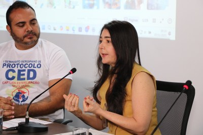 Mulher sentada à mesa fala ao microfone enquanto gesticula com as mãos. Ao lado dela, um homem observa a fala. Ambos estão posicionados diante de uma tela de projeção ao fundo.