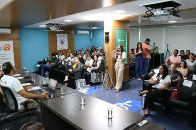Auditório com público sentado assistindo a uma mesa de apresentações do MPF. Quatro pessoas compõem a mesa ao fundo, enquanto participantes, fotógrafos e cinegrafistas estão distribuídos pela sala. Alguns banners sobre ações voltadas à causa animal estão expostos nas paredes.