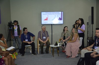 Quatro debatedores estão sentados em uma roda de conversa, com uma moderadora falando em pé ao lado. Um monitor exibe um gráfico sobre "Extremos Climáticos" no Brasil.