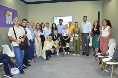 Uma foto de grupo posada com cerca de 18 participantes de um evento, em um espaço de debate. Eles estão em pé, olhando para a câmera, com um monitor e um banner roxo com a frase "SOU GUARDIÃO DO FUTURO" ao fundo.