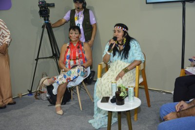 Duas mulheres indígenas, com pinturas faciais e adereços, sentadas durante um debate. A mulher da direita fala ao microfone, enquanto a da esquerda observa. Um cinegrafista as grava ao fundo.