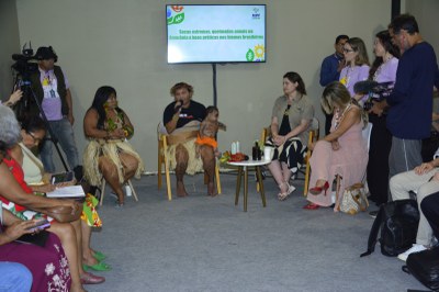 Duas mulheres indígenas em trajes tradicionais (saia de palha), sentadas, participam do debate. Uma delas segura um bebê e a outra fala ao microfone.