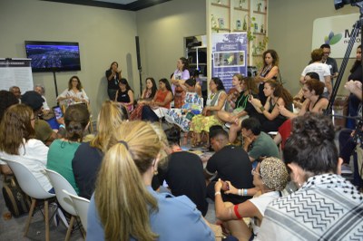 Uma roda de conversa com cerca de 30 pessoas, a maioria mulheres, em um ambiente fechado. Algumas estão sentadas em cadeiras e outras no chão, prestando atenção em quem fala em pé ao fundo.