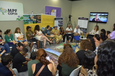 
Uma roda de conversa com cerca de 30 pessoas em um evento, no estande do MPF na  COP30. Um grupo está sentado em cadeiras no centro, e os demais formam um semicírculo em volta, sentados no chão ou em cadeiras brancas. Um monitor exibe uma pessoa falando.